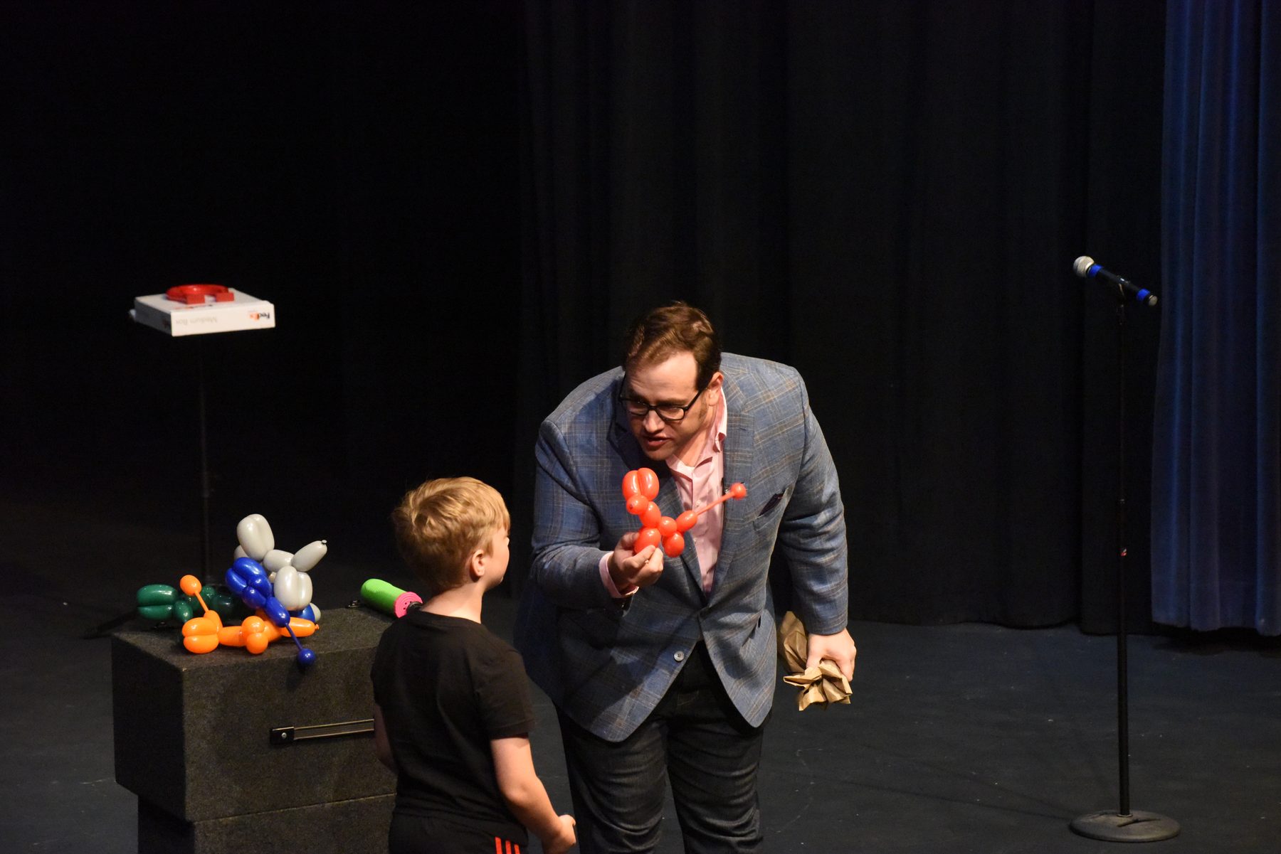 Scott Green handing a balloon animal to a young volunteer on stage