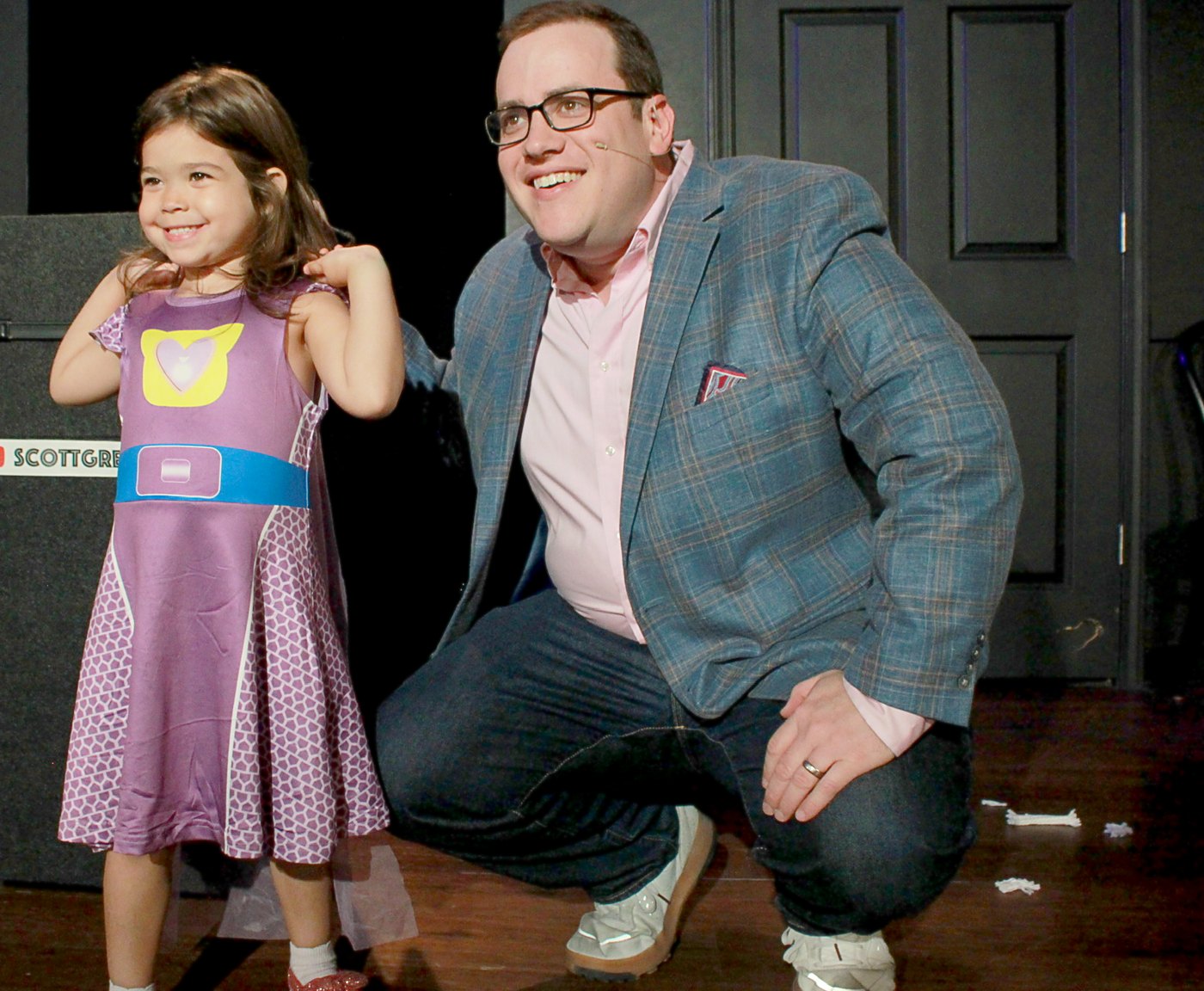 Scott Green crouched beside a beaming young volunteer on stage at iO Theater